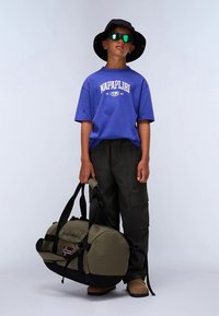 Žalia ir juoda kelioninė kuprinė su šoninėmis rankenomis ir vėliavos detale. Modelis dėvi mėlyną marškinėlį, juodą kibiro formos kepurę ir cargo stiliaus kelnes.