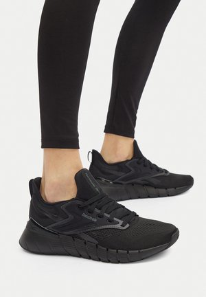 Füße in schwarzen Reebok-Laufschuhen und schwarzen Leggings, dargestellt in stehender Pose vor einem einfarbigen weißen Hintergrund.