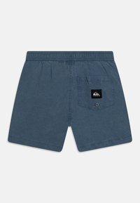 Modré šortky vyrobené z měkkého, texturovaného materiálu, s elastickým pasem a zadní kapsou s černou nášivkou s logem.