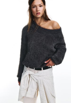 Vrouw draagt een donker, pluizig off-shoulder trui, lichte broek met een gestreept overhemd om de taille geknoopt, staande tegen een witte achtergrond.