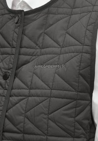 Črna prešita telovnik z diamantnim vzorcem, spredaj z zaponko na pritisni gumb in sivim vezenim logotipom "KARL LAGERFELD." Vidni so detajli teksturirane površine.