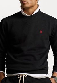 Svart crewneck sweatshirt i ett mjukt material, med en röd polo-logotyp på bröstet, ihop med en vit skjorta med krage under.