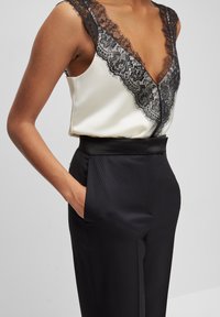 Hedvábný top na ramínka s černou krajkovou lemovkou a hlubokým výstřihem do V, doplněný o černé kalhoty na míru s hladkým pasem a bočními kapsami.