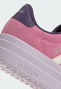 Gros plan d'une basket Adidas en daim rose avec languette de talon violette et semelle violet clair avec logo Adidas embossé.