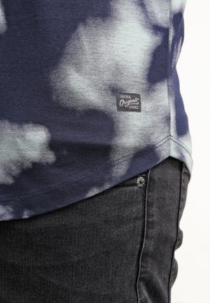 Gros plan sur l'ourlet d'un t-shirt tie-dye bleu et blanc avec l'étiquette "Jack & Jones Originals" au-dessus de la poche d'un jean en denim noir.