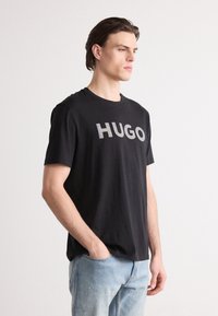 Melnas kokvilnas t-krekls ar īsām piedurknēm, kurā krūtīs ir atstarojošā burti "HUGO". Valkāts ar gaišām džinsu biksēm.