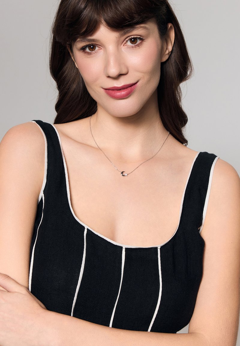 Srebrny naszyjnik z zawieszką w kształcie półksiężyca. Modelka ma na sobie czarną bluzkę w paski z białymi akcentami, prezentującą gładki, teksturowany materiał.