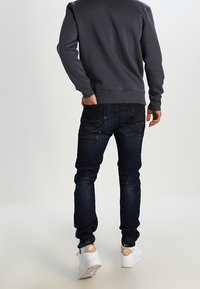 Jeans in denim blu scuro con una vestibilità slim, caratterizzati da un sottile effetto di sbiancatura, abbinati a sneaker bianche e a una felpa grigia.