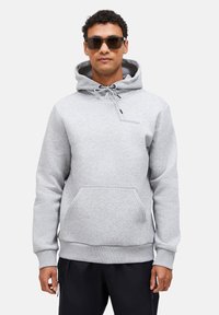 Felpa grigia con cappuccio, dettagli con stringhe e tasca frontale. Presenta polsini e orlo a costine. Il modello indossa occhiali da sole e pantaloni scuri.