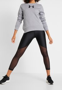 Grijze sweatshirt met zwart logo, gecombineerd met zwarte legging met doorschijnende panelen en gladde textuur. Zwarte sportschoenen maken de look af.