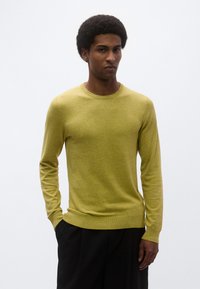 Κίτρινο πουλόβερ με στρογγυλή λαιμόκοψη σε απαλό πλεκτό ύφασμα, με μακριά μανίκια και ριμπ μανσέτες, συνδυασμένο με μαύρο παντελόνι.