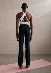 Biały sportowy stanik z krzyżowanymi ramiączkami i otwartym tyłem, w zestawie z czarnymi spódniczkami jogowymi. Gładka tekstura na wszystkich ubraniach, stojąc na różowym macie.