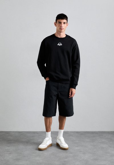 Svart sweatshirt med en liten grafik, kombinerad med svarta shorts och vita sneakers med en beige sula. Modellen står med händerna i fickorna.
