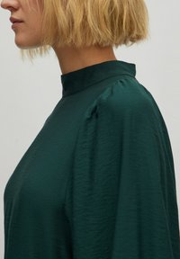 Dunkelgrüne, strukturierte Bluse mit hohem Kragen und puffärmel, die einen geschmeidigen Stofffall und eine taillierte Silhouette aufweist.