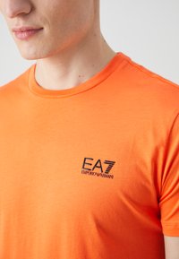 Oranžs kokvilnas T-krekls ar apaļu kakla izgriezumu un īsām piedurknēm, ar melnu EA7 Emporio Armani logo kreisajā krūtī.