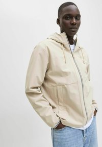 Chaqueta ligera beige con capucha, con cremallera frontal, dos bolsillos laterales y puños a rayas en contraste; combinada con una camiseta blanca y jeans.