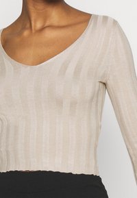 Haut en maille côtelée beige à manches longues avec un décolleté en V profond et un ourlet festonné, présentant une texture douce et un motif à rayures verticales.
