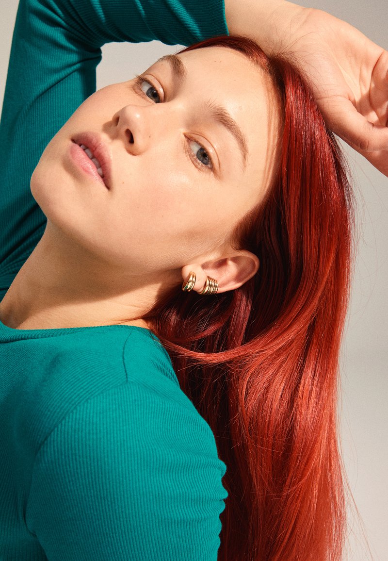 Goldene Creolen an einem Modell mit langen, glatten roten Haaren, das ein taublauses, geripptes Langarmshirt trägt und vor einem neutralen Hintergrund posiert.