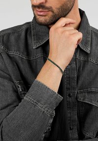 Braccialetto di perline con perline verde scuro e nere, design a cordoncino; indossato al polso sopra una camicia di jeans grigia con dettagli a bottoni.