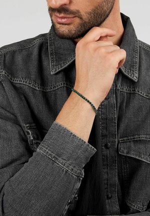 Braccialetto di perline con perline verde scuro e nere, design a cordoncino; indossato al polso sopra una camicia di jeans grigia con dettagli a bottoni.