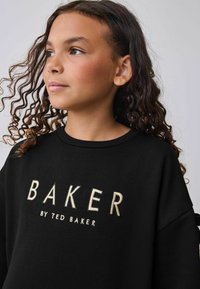 Mustapaita, jossa on "BAKER"-logo vaaleanbeigeillä kirjontatöillä. Ominaisuuksiin kuuluu pyöreä pääntie ja pitkät hihat, ja paita on muotoiltu rentoon istuvuuteen.