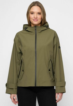 Vrouw die een olijfgroene gewatteerde jas met capuchon draagt, voorzien van een ritssluiting aan de voorkant, zakken met kleppen en knopen, en verstelbare polsbandjes, staand tegen een witte achtergrond.