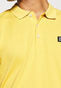 Gros plan sur une personne portant un polo jaune avec col, trois boutons et un petit écusson noir et blanc sur la poitrine.