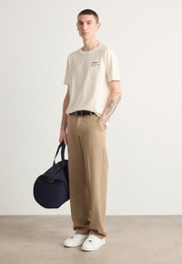 Beige katoenen t-shirt met een klein zwart logo, tan kleurige broeken, witte sneakers en een ronde marineblauwe tas met een gouden accent. Model staat tegen een effen achtergrond.