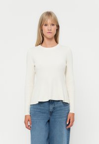 Light cream long-sleeve top with a fitted bodice and flared hem, made from soft, ribbed fabric. Paired with high-waisted blue jeans.