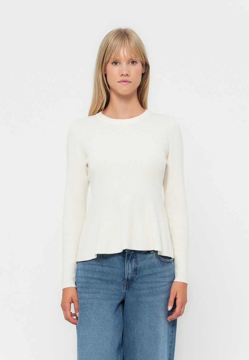 Light cream long-sleeve top with a fitted bodice and flared hem, made from soft, ribbed fabric. Paired with high-waisted blue jeans.
