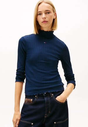 Marineblauw geribd coltruitje met geplooide kraag, lange mouwen en subtiel logodetail op de borst, gecombineerd met donkerdenim broek.