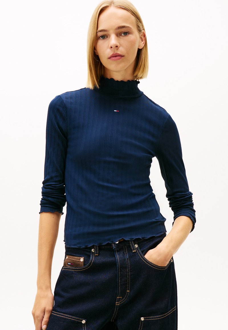 Ναυτικό μπλουζάκι με πλεκτό ζιβάγκο, γυαλιστερό γιακά με βολάν, μακρυά μανίκια και διακριτική λεπτομέρεια λογότυπου στο στήθος, συνδυασμένο με σκουρόχρωμο τζιν παντελόνι.