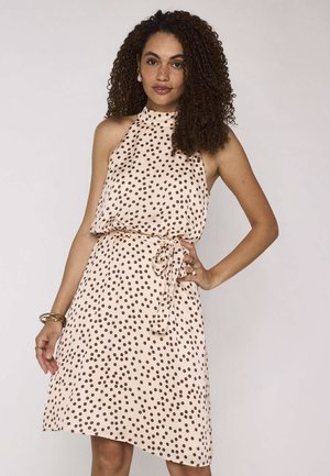 Vrouw met krullend haar die een mouwloze crèmekleurige jurk met bruine polkadots en een gestrikte tailleband draagt, poseren met één hand op de heup.