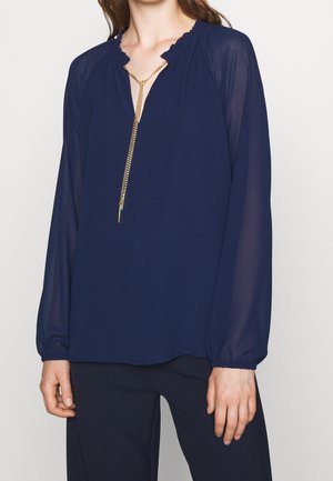 Vrouw draagt een marineblauwe blouse met lange, doorschijnende mouwen en een gouden ketting, gecombineerd met een donkere broek.