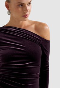 Violet, bezplecu kleita no mīksta, spīdīga auduma ar plūstošām detaļām un garām piedurknēm. Dekoratīvas auskari papildina šo izskatu.