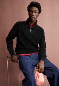 Junger Mann sitzt, trägt einen schwarzen Reißverschluss-Pullover, blaue Jeans und hält eine rosa Mütze vor einem gedeckten rosa Hintergrund.