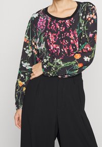 Blumenbluse mit langen Ärmeln und rundem Ausschnitt. Sie zeigt lebendige rosa, orange und weiße Blumen auf einem schwarzen Hintergrund. Faltenmuster im Oberteil.