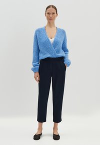Blauwe gebreide cardigan met een diepe V-hals, gecombineerd met zwarte taps toelopende broek en zwarte flats. De outfit heeft een gestructureerd garen.
