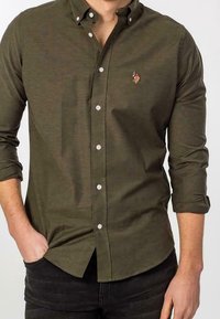 Olive green button-up shirt with a collar, featuring white buttons and a small embroidered detail on the chest, long sleeves rolled at the cuffs.