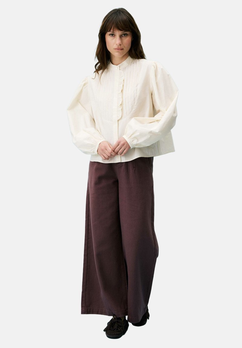 Femme portant un chemisier crème à manches bouffantes et un pantalon large marron, debout les mains jointes devant sur un fond uni.