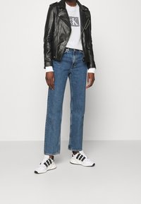 Musta nahkainen moottoripyörätakki, valkoinen graafinen huppari, siniset suorat farkut, valkoiset ja mustat sneakerit raidallisilla yksityiskohdilla.