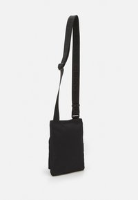 Sac messager en tissu noir avec une bandoulière réglable unique, présentant une texture lisse et un logo embossé discret sur la bandoulière.