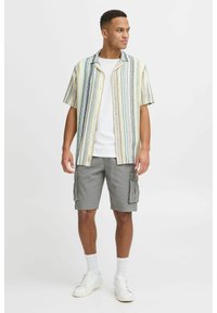 Kurzärmliges gestreiftes Hemd in Gelb, Blau und Weiß, kombiniert mit grauen Cargo-Shorts. Weiße Sneakers und Socken runden das Outfit ab.