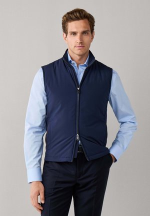 Homme portant un gilet zippé bleu marine sur une chemise bleu clair et un pantalon sombre, debout avec une main dans la poche devant un fond uni.