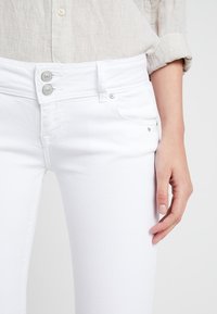 Jeans de mezclilla blanca con una textura suave, cierre de dos botones, bolsillo delantero y acentos metálicos cerca del bolsillo.