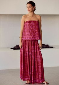 Robe maxi sans bretelles en bordeaux avec des motifs rosesintriqués. Tissu fluide, taille froncée et coupe décontractée. Associée à des sandales simples.