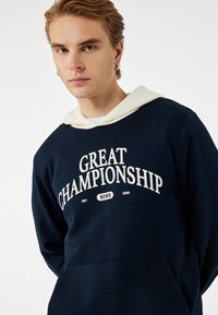 Sudadera navy con cordón blanco y texto impreso "GRAN CAMPEONATO". Confeccionada en una tela suave, cuenta con un bolsillo frontal y una capucha de contraste.