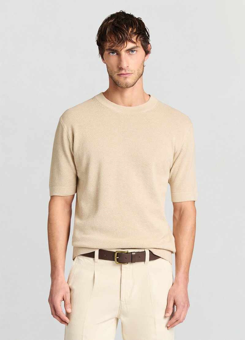 Pull beige en maille à manches courtes avec un col rond, motif texturé, porté avec un pantalon de couleur claire et une ceinture marron.