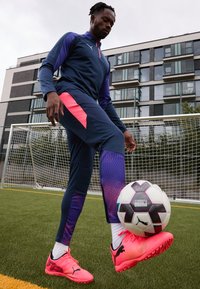 Puma sportinė apranga, apimanti tamsiai mėlyną marškinėlį su rožinėmis detalėmis ir tekstūruotas kelnes. Ryškiai rožiniai futbolo batai, juodas ir baltas futbolo kamuolys po kojomis.