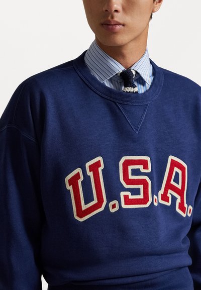 Sweatshirt marine avec "U.S.A." en broderie rouge et blanche sur la poitrine. Doté d'un col rond et d'un tissu texturé. Chemise rayée en dessous.